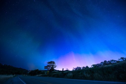 A serene night sky filled with stars, featuring a gradient of blue and purple hues above a quiet road and silhouette of trees.