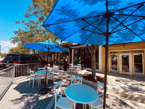 Outdoor café scene with blue umbrellas, white tables, and chairs, set against a sunny street backdrop.