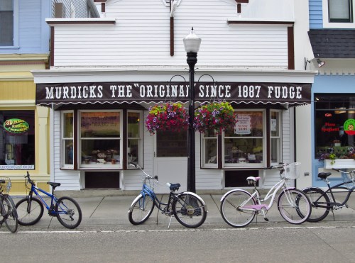 A quaint fudge shop with a sign reading "Murdicks The Original Since 1887" and colorful flower baskets outside. Bicycles parked nearby.