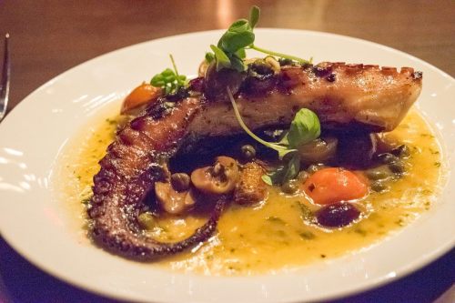 Grilled octopus served on a plate with vegetables and a flavorful sauce, garnished with fresh herbs.