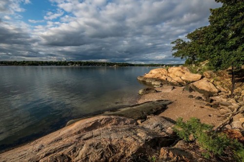 A serene lakeside view with rocky shorelines, lush greenery, and a cloudy sky reflecting on the calm water.