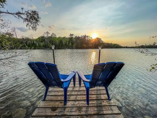 Two blue chairs on a wooden dock overlooking a calm lake at sunset, surrounded by trees.