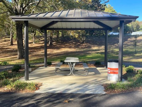 A shaded picnic area with a metal roof, table, and trash can in a park setting surrounded by trees.