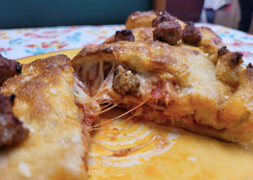 A close-up of a cheesy stuffed pizza with sausage, showing melted cheese and a golden crust on a colorful tablecloth.