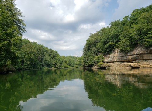 Serene river scene surrounded by lush green trees and rocky cliffs under a partly cloudy sky.