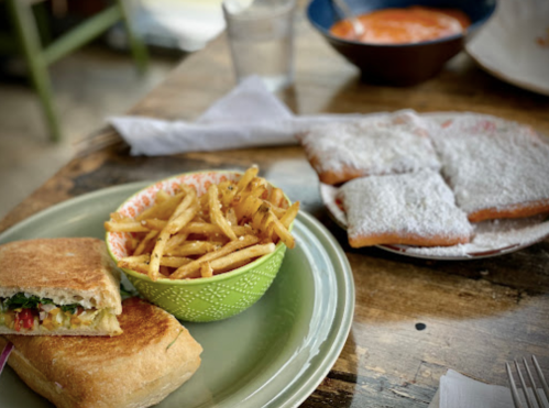 A plate with a sandwich, a bowl of fries, and a plate of powdered sugar-covered beignets on a wooden table.