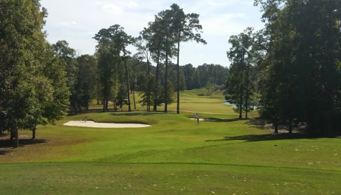 A scenic golf course with a sand trap, trees, and a player in the distance under a clear blue sky.
