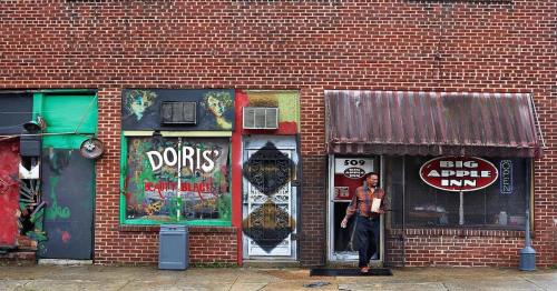 A man stands at the entrance of a brick building with colorful storefronts, including "Doris' Beauty Black" and "Big Apple Inn."