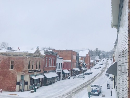 Snow-covered street in a small town, featuring historic buildings and parked cars under a gray winter sky.