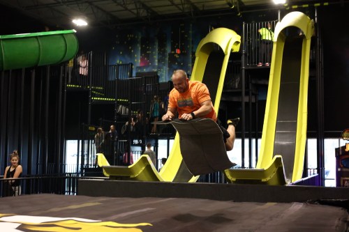 A person in an orange shirt jumps off a slide at an indoor amusement park, preparing to land on a soft surface below.