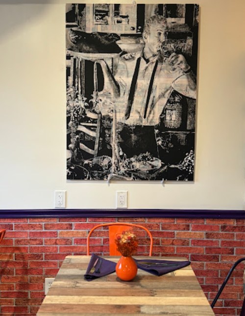 A black-and-white artwork of a man serving food hangs above a rustic wooden table with orange chairs and a vase of flowers.