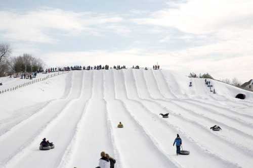 A snowy hill with people tubing down, surrounded by trees and a cloudy sky.