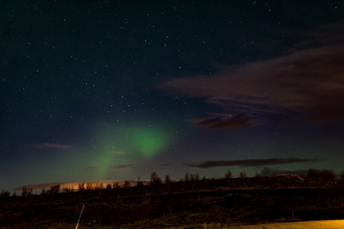 A night sky filled with stars and a faint green aurora over a dark landscape with clouds.
