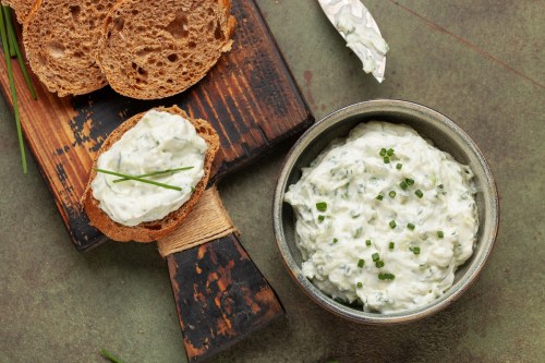 A bowl of creamy herb spread with chives, served with slices of brown bread on a wooden board.