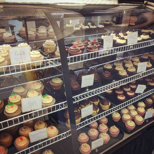 A display case filled with various colorful cupcakes, each labeled with their flavors.