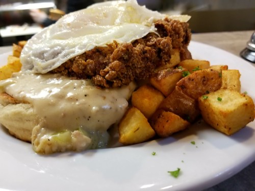 A plate of fried chicken topped with a fried egg, served over biscuits and gravy, alongside crispy potato cubes.