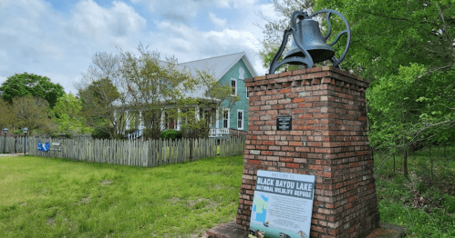 A brick monument with a bell and sign stands in front of a green house, surrounded by trees and a wooden fence.