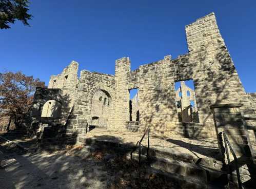 Ruins of a stone building with arched windows and doorways, surrounded by trees under a clear blue sky.