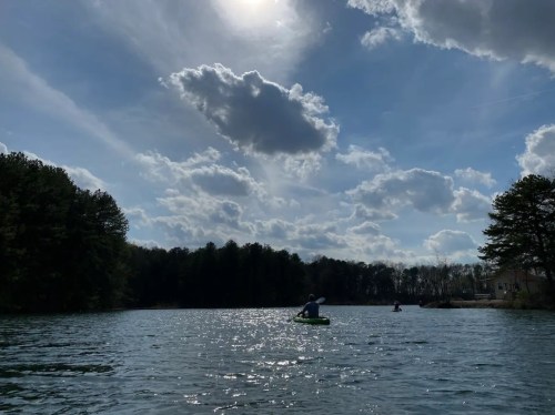 A serene lake scene with a person kayaking under a bright sky filled with clouds and sunlight reflecting on the water.