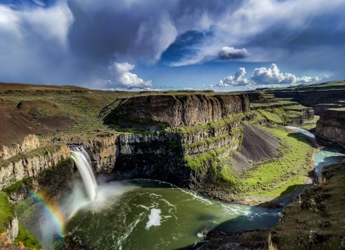 A stunning waterfall cascades into a river, surrounded by lush green cliffs and a vibrant rainbow under a cloudy sky.