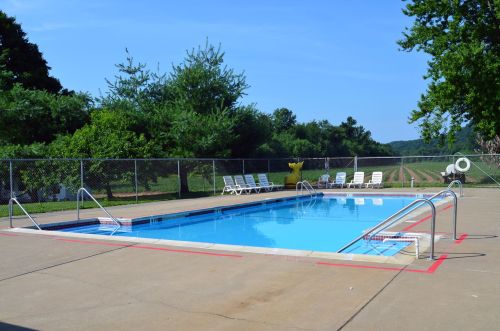 A clear blue swimming pool surrounded by lounge chairs and greenery, with a fence and fields in the background.