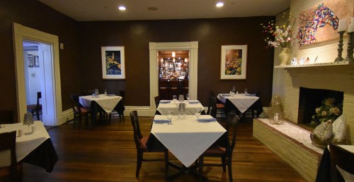 A cozy restaurant interior with white tablecloths, wooden floors, and a bar visible through an archway.