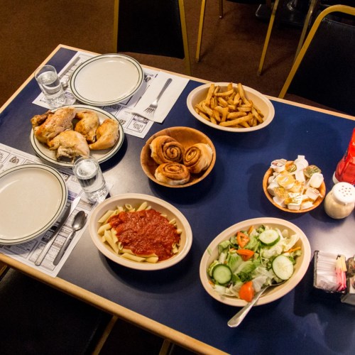A table set with plates of chicken, pasta with sauce, salad, fries, cinnamon rolls, and empty plates.