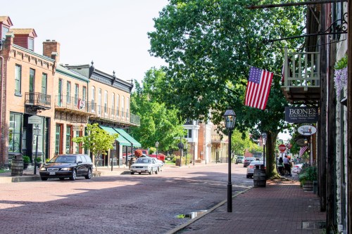 A charming street lined with historic buildings, American flags, and trees, featuring cobblestone pavement and parked cars.