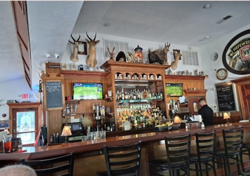 A cozy bar with wooden decor, mounted animal heads, two TVs showing sports, and a well-stocked liquor shelf.