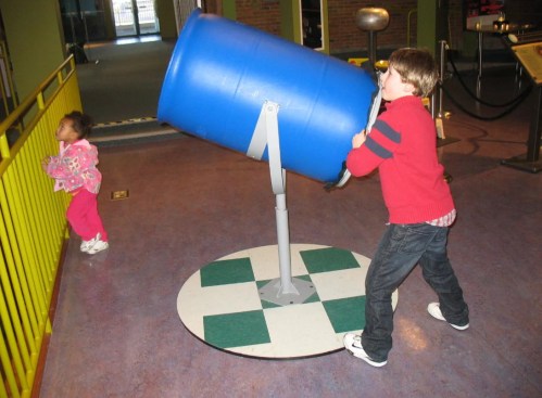 A child in a red sweater lifts a blue barrel at a science exhibit, while a toddler in pink watches nearby.