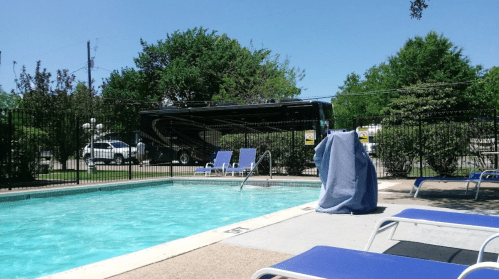 A clear blue pool surrounded by lounge chairs, with an RV parked nearby and greenery in the background.