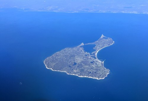 Aerial view of a small island surrounded by blue water, featuring green land and a coastal outline.