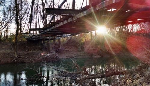 An old, rusty bridge spans a calm river, with sunlight filtering through trees, creating a serene atmosphere.