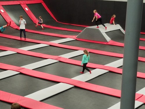 Children jumping and playing on trampolines in a trampoline park with red and gray surfaces.