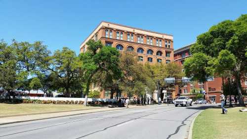 A sunny street view of a brick building surrounded by trees and greenery, with people walking nearby.