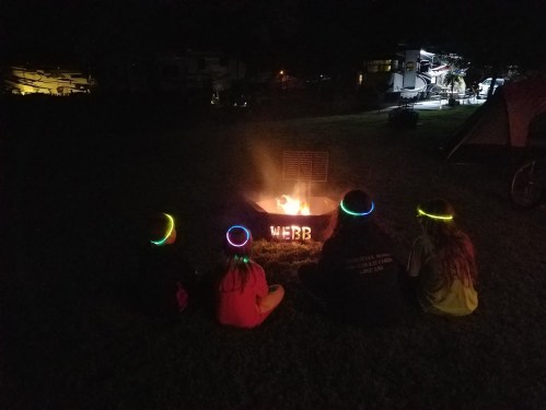Four people sit around a campfire at night, wearing glowing bracelets, with "WEBB" illuminated in the fire pit.