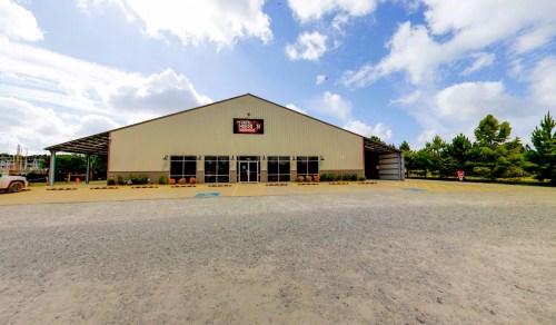 A large metal building with a sign, surrounded by gravel and trees, under a partly cloudy sky.