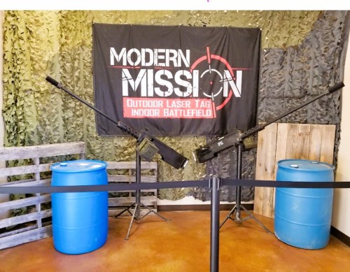 A laser tag arena featuring two mounted laser guns, blue barrels, and a camouflage backdrop with a "Modern Mission" banner.
