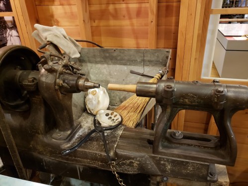 An old woodworking lathe with tools and a broom on a wooden workbench in a rustic setting.