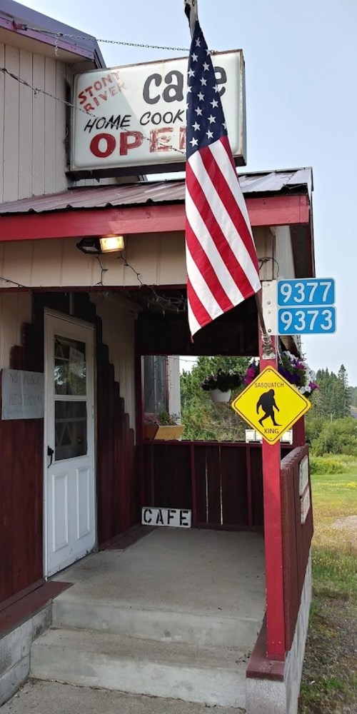 A rustic café entrance with an American flag, a "Sasquatch Crossing" sign, and an "Open" sign above the door.