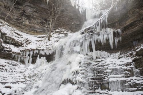 A frozen waterfall cascading down rocky cliffs, surrounded by snow and icicles in a winter landscape.