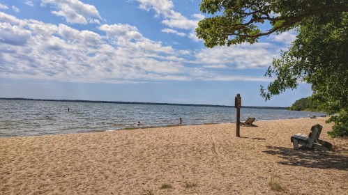 A sandy beach by a calm lake, with a few people swimming and trees lining the shore under a partly cloudy sky.