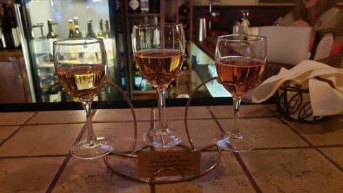Three wine glasses with different colored wines on a bar counter, displayed in a metal holder.