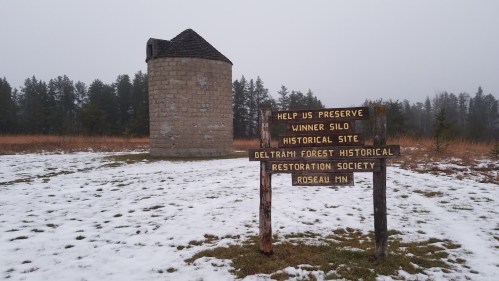 A stone silo with a sign about its historical preservation, surrounded by snow and trees in a foggy landscape.