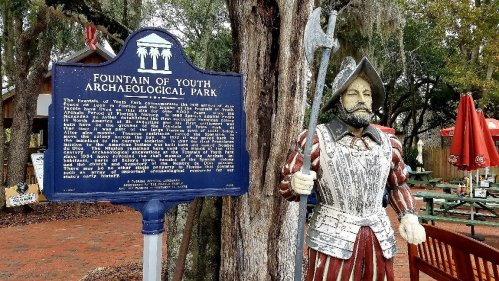 A historical marker for Fountain of Youth Archaeological Park next to a statue of a Spanish explorer.