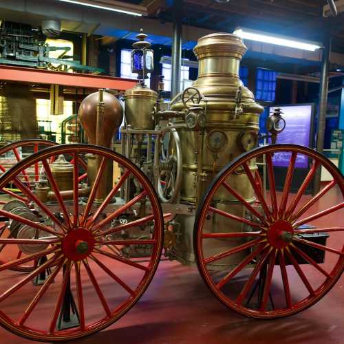 A vintage steam fire engine with large red wheels, displayed in a museum setting with industrial background.