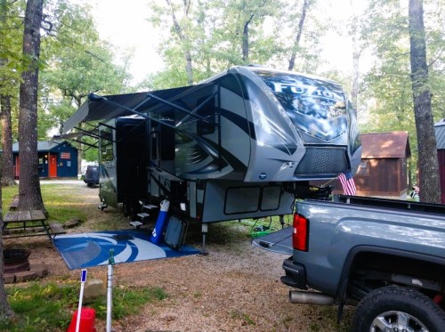 A large RV parked in a wooded area, with an awning extended and a pickup truck nearby.