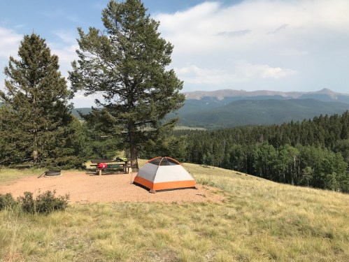 A tent set up on a grassy hillside, surrounded by trees, with mountains in the background under a partly cloudy sky.