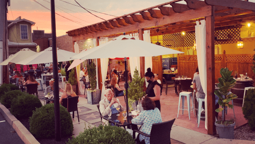 Outdoor dining area with tables under umbrellas, surrounded by greenery, at sunset. People enjoying their meals.