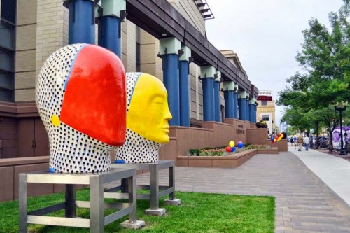 Colorful abstract sculptures of heads in red and yellow, placed outside a modern building with blue columns.